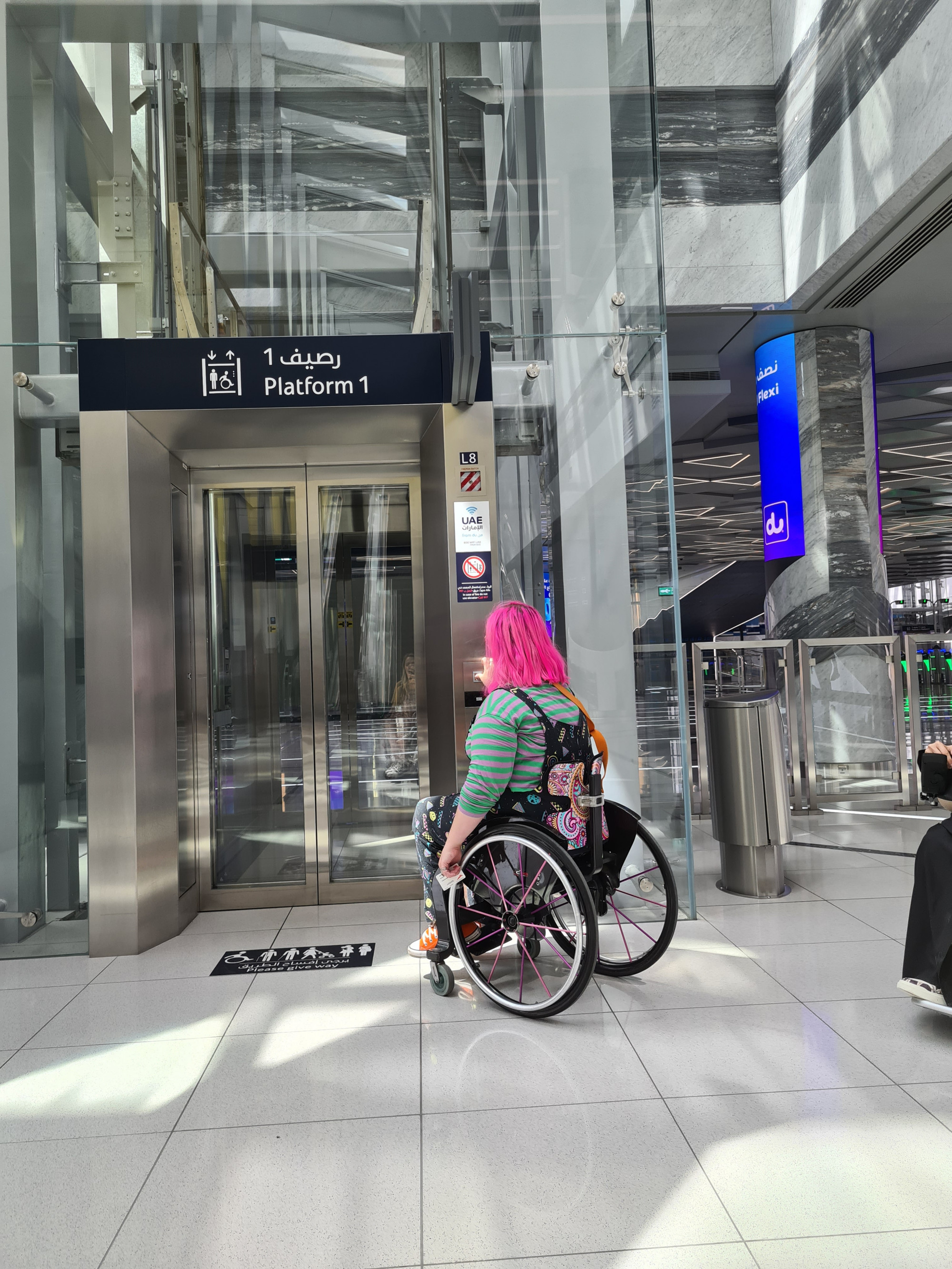 A photograph of Emily Yates of Mima  in her wheelchair in front of the doors to a large steel and glass elevator, with a sign saying 