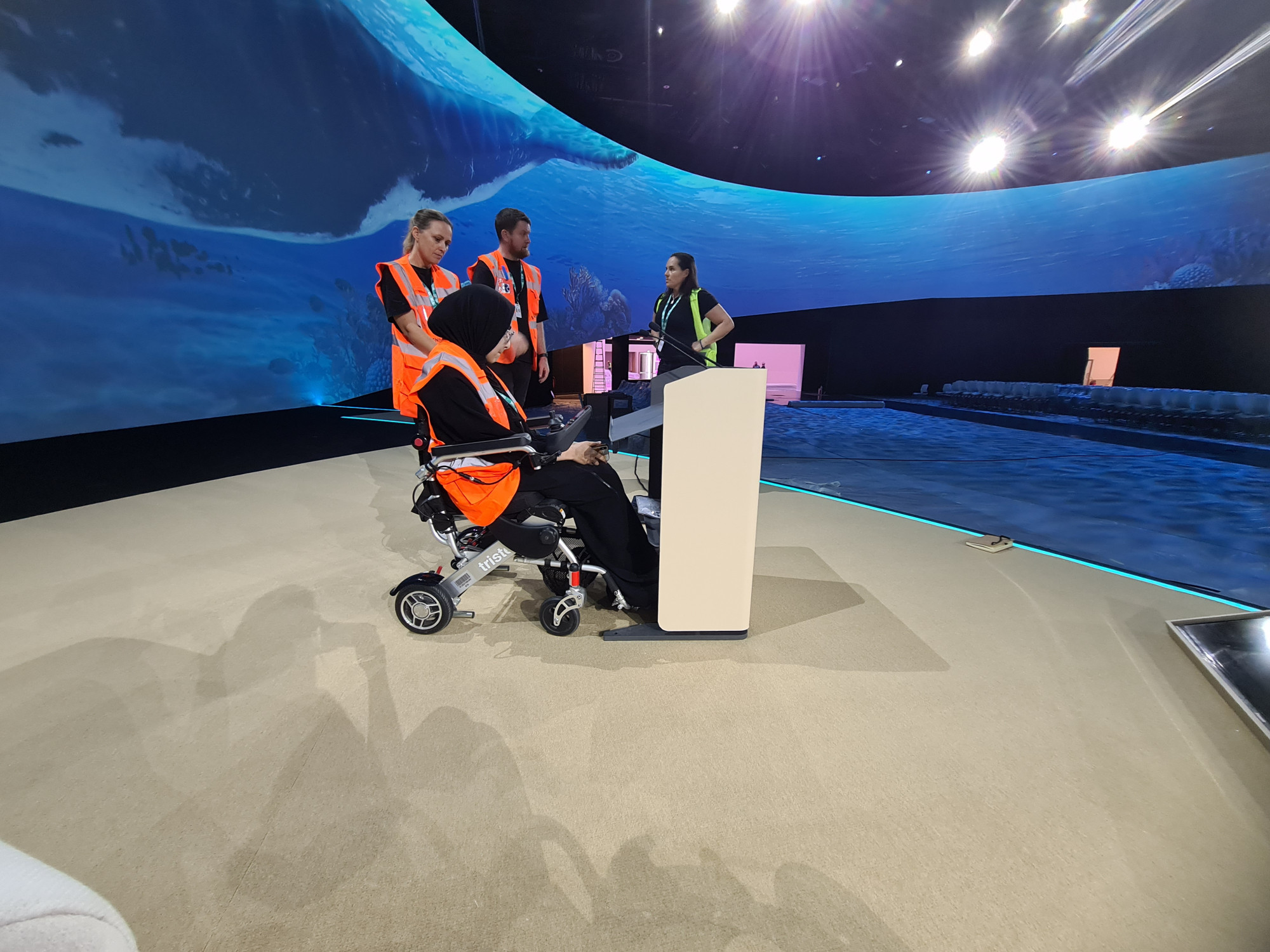 A photograph showing access consultant and powerchair user Fatma Aljassim testing a height adjustable lectern on the COP28 blue zone stage. Three members of the COP28 design team are stood behind Fatma having a conversation.