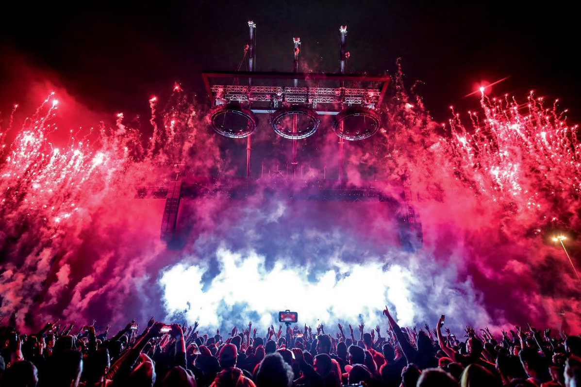 An outdoor festival scene, with dark sky, stage and red and purple flares exploding around a dancing crowd.
