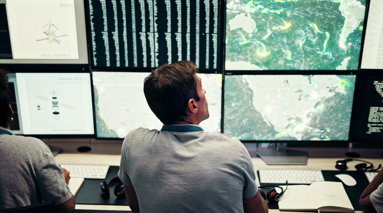 A Control room operator sits in front of a wall of TV screens  monitoring weather and satellite maps.