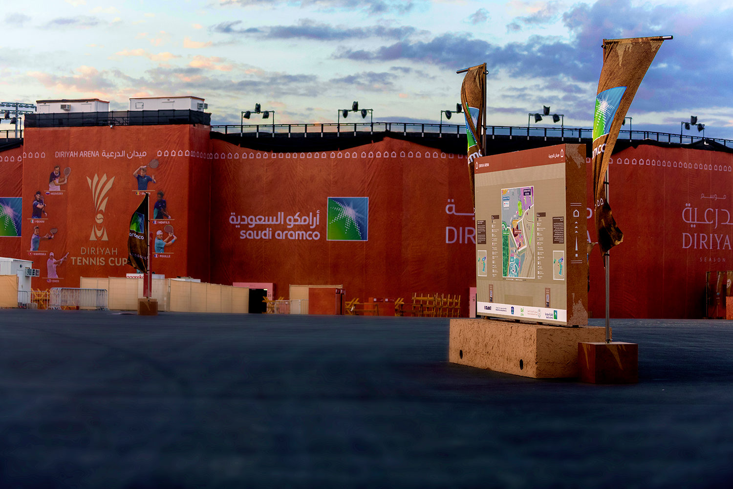 Mima's wayfinding map at Diriyah, with colourful elements on a brown background and installed on a brown podium, is to the right of the image. Orange advertisements line the wall in the background, and the sky is blue.