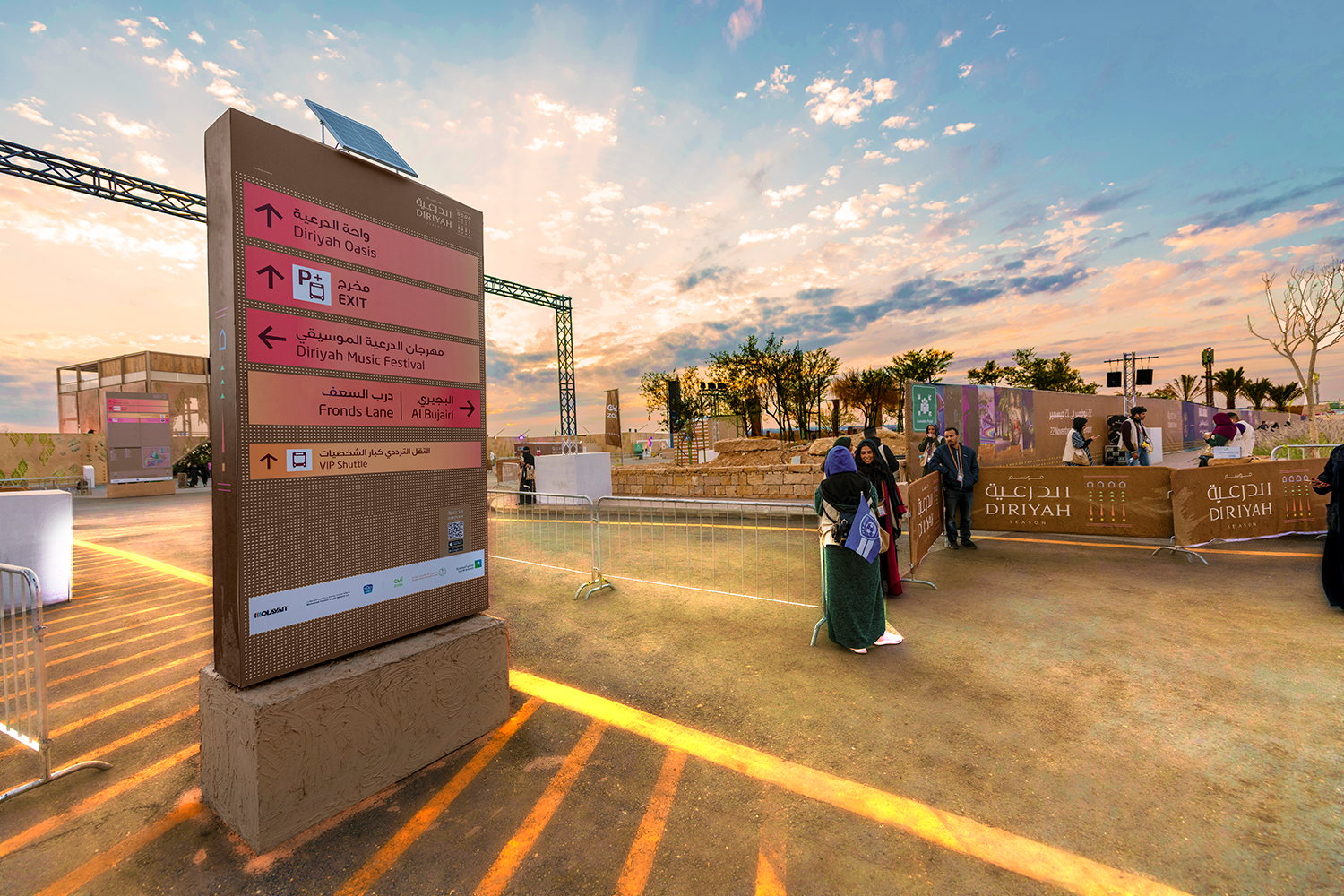 Mima's red and orange wayfinding signage, on a brown background, in place at Diriyah. Visitors and in the background and the sky is blue.