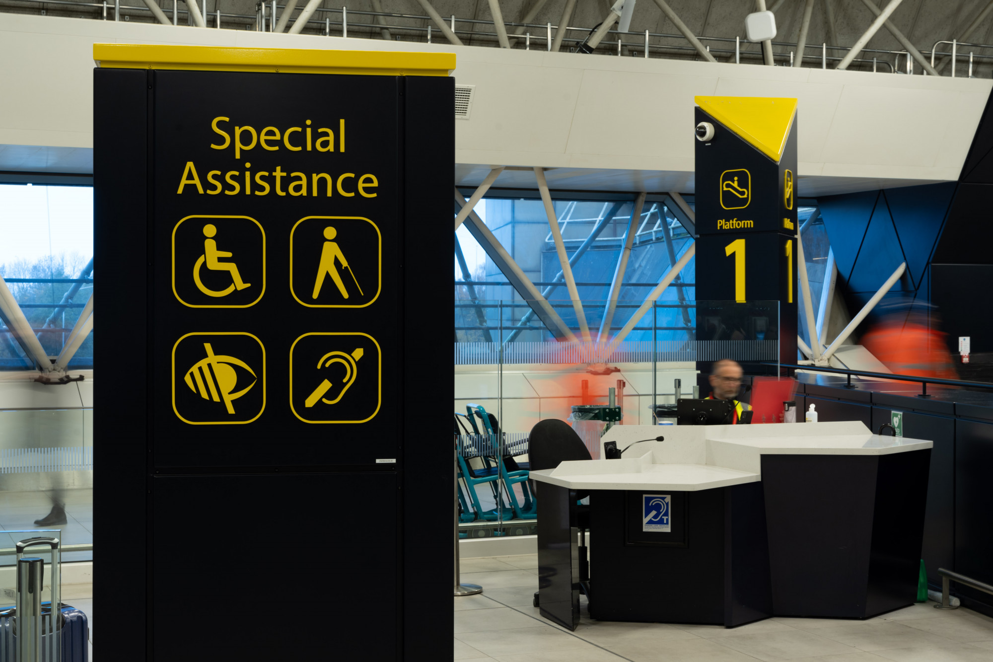 A photo of the black wayfinding totem designed specifically for Gatwick Airport station with yellow icons depicting special assistance and access impairments, with the accessible customer inquiry desk in the background.