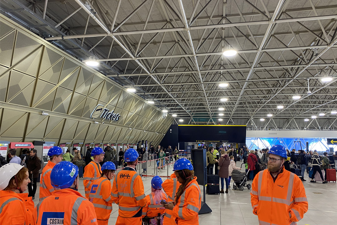 The Mima team on site at Gatwick airport train station wearing high vis orange jackets and blue hard hats. Passengers are moving around the ticket hall with suitcases.
