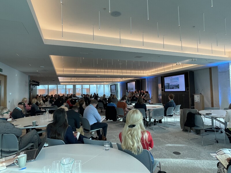 A photo taken from the event and the right-hand side of a large room filled with circular tables full of conference delegates facing two large screens while a panel debate takes place.