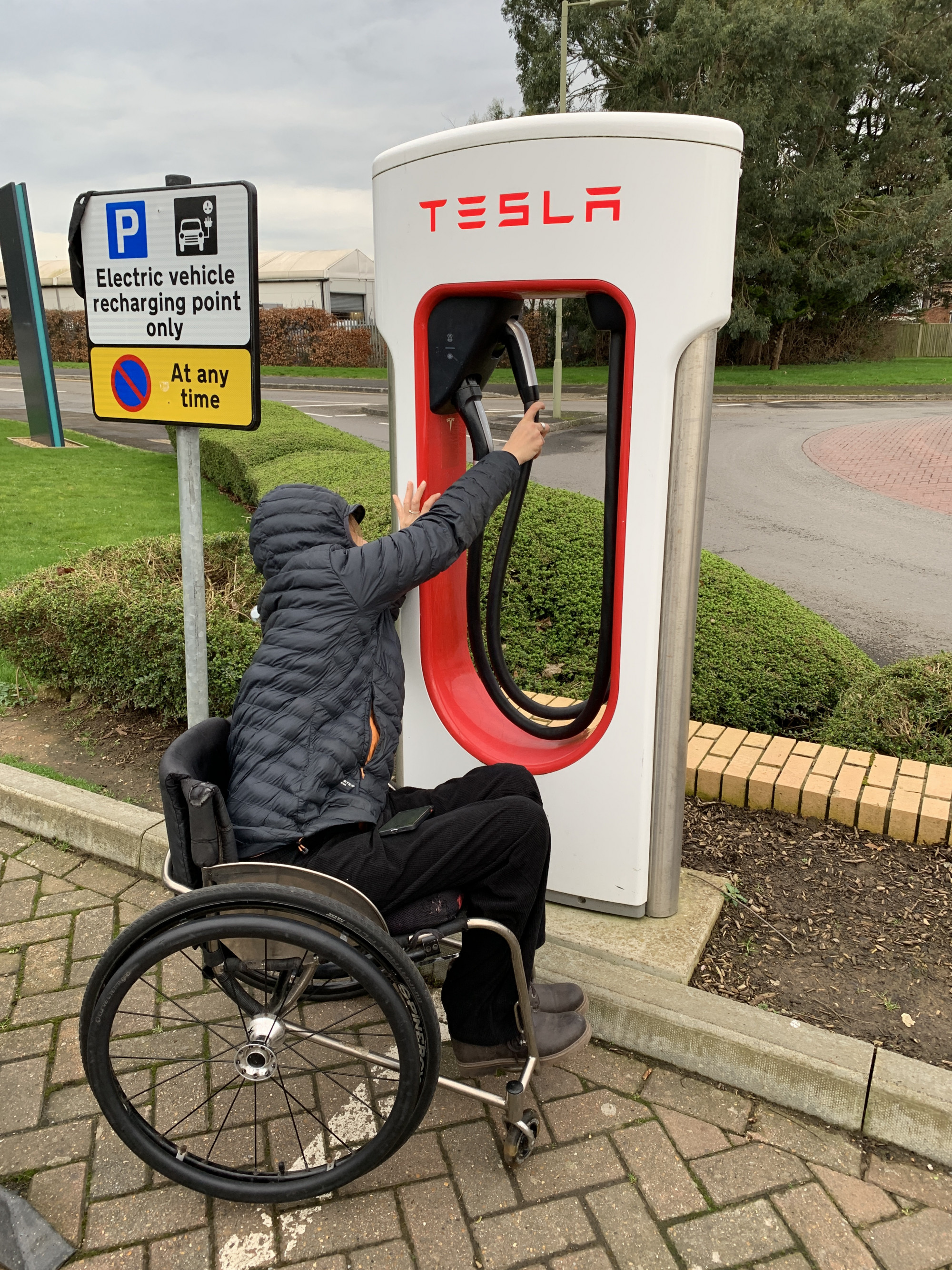 A photo of our Senior Access consultant Tash Davies reaching for a Tesla charger positioned above shoulder height, at the limit of her range of motion, making it difficult to access.