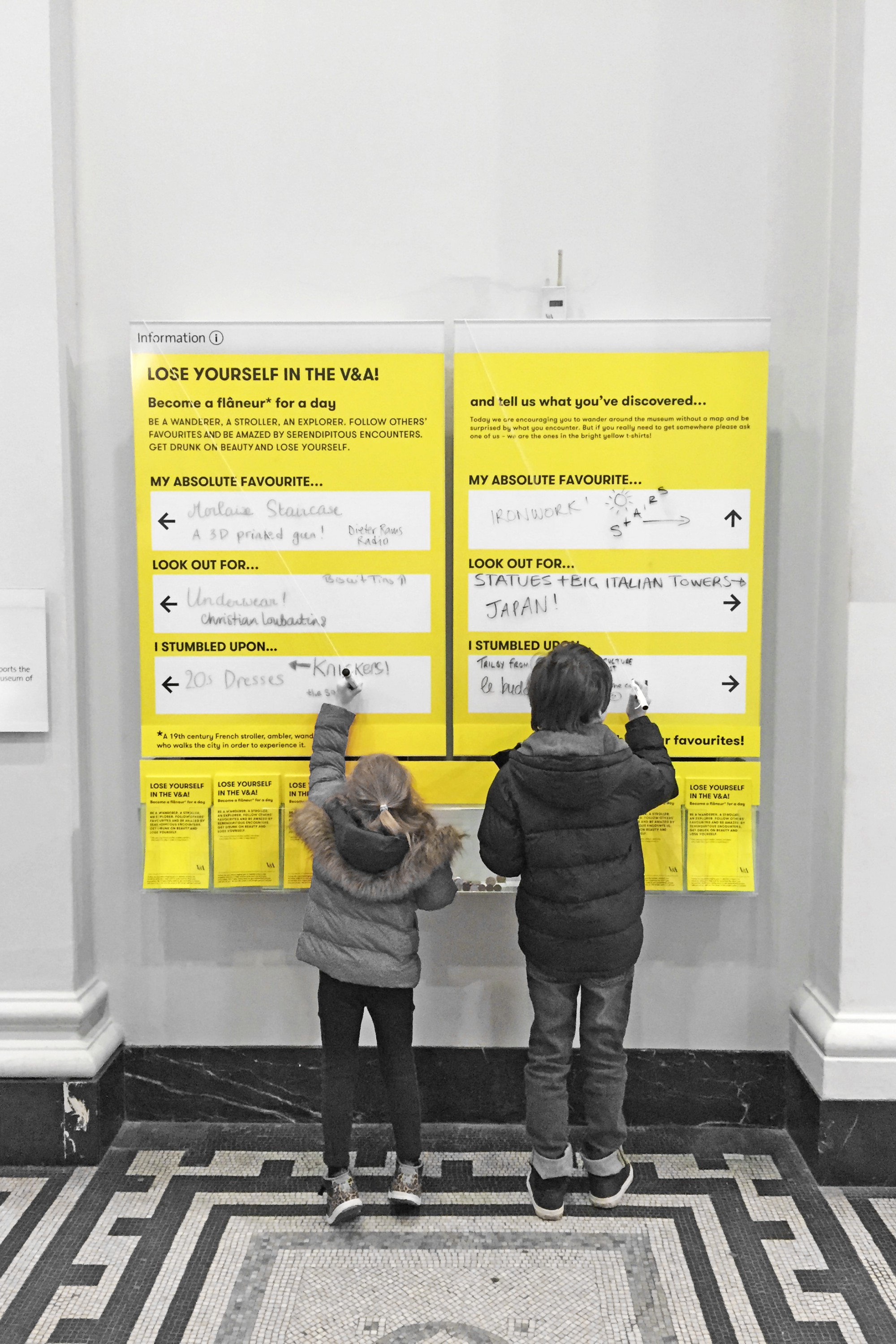 Two children stand in front of a yellow activity board at the V&A museum and write on it with marker pens. The board  asks visitors to share what they've discovered during their visit, and encourages others to lose themselves whilst exploring.