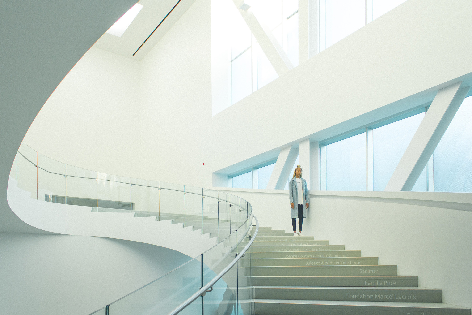 A woman stands still on a wide contemporary spiral sweeping staircase in a museum or open space, with large windows on the right side letting in light. The edge of each stair has subtle text engraved into it.