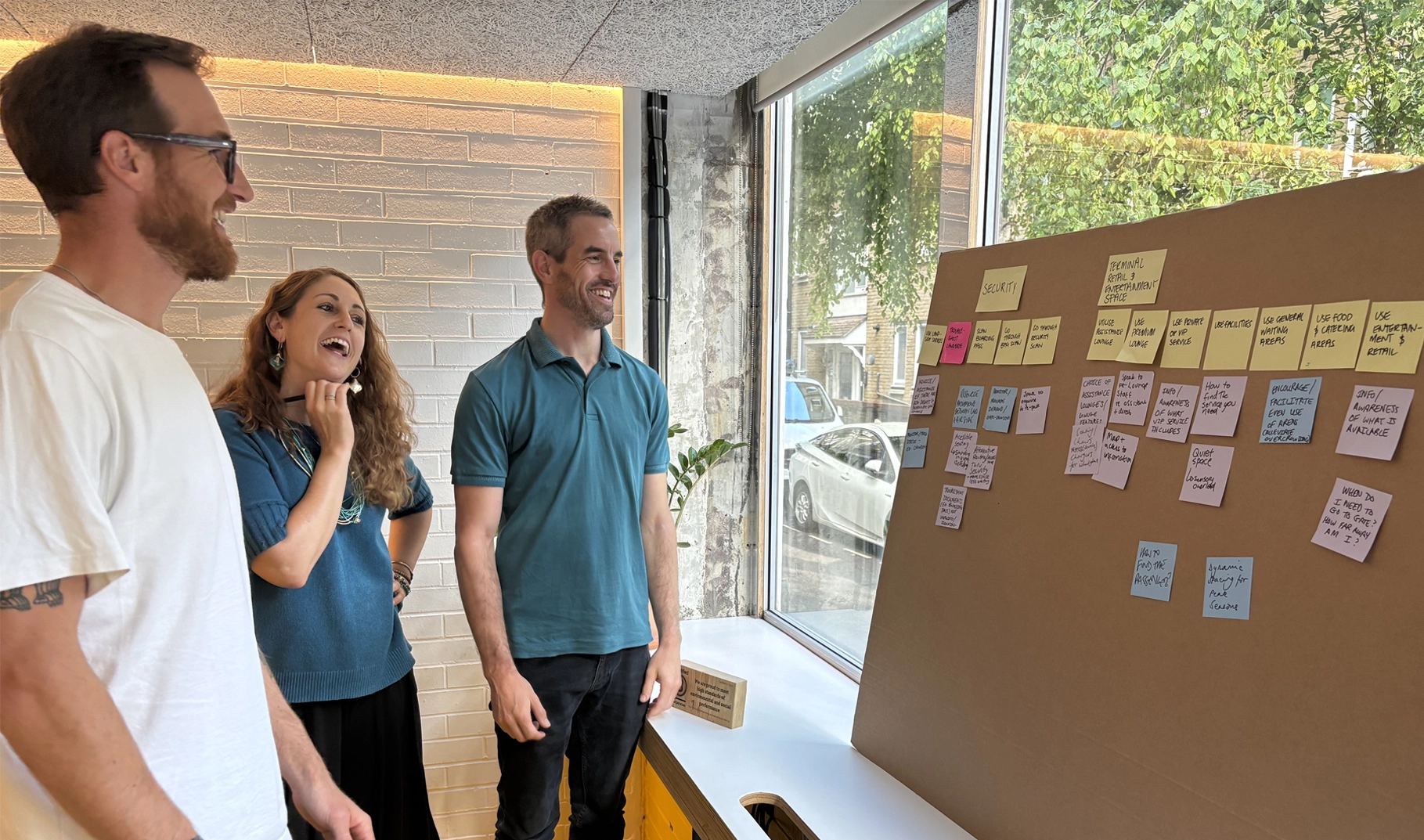 Three people stand in front of a large brown board with post it notes on. They are talking and writing notes on the board.