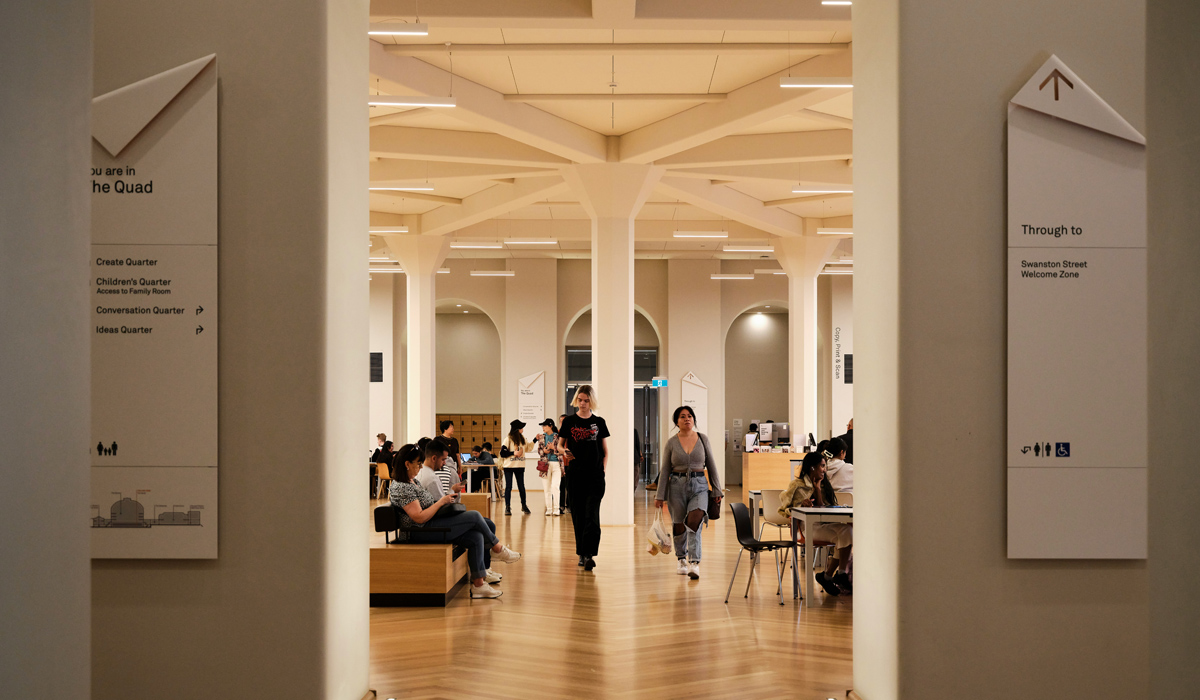 Looking through an archway in a contemporary museum, with directional signage either side of the arch. Beyond, people are sat down, with two people walking in the centre.