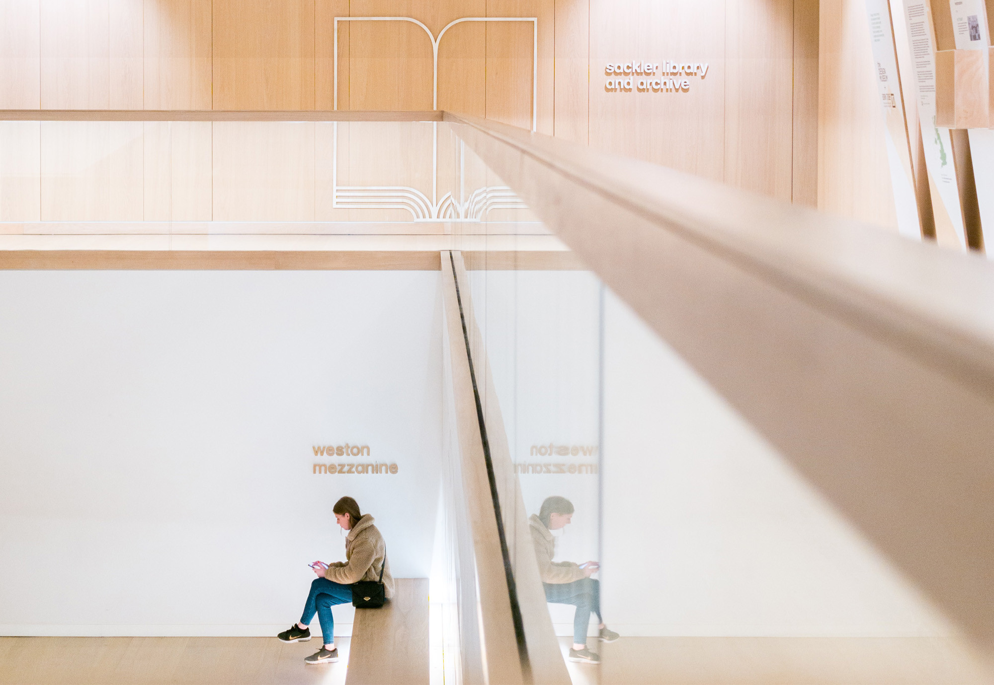 A museum visitor sits on a bench on a lower open plan level in a contemporary museum, looking down at their phone. Above them another floor of the museum with a glass walled barrier, and light wooden walls.