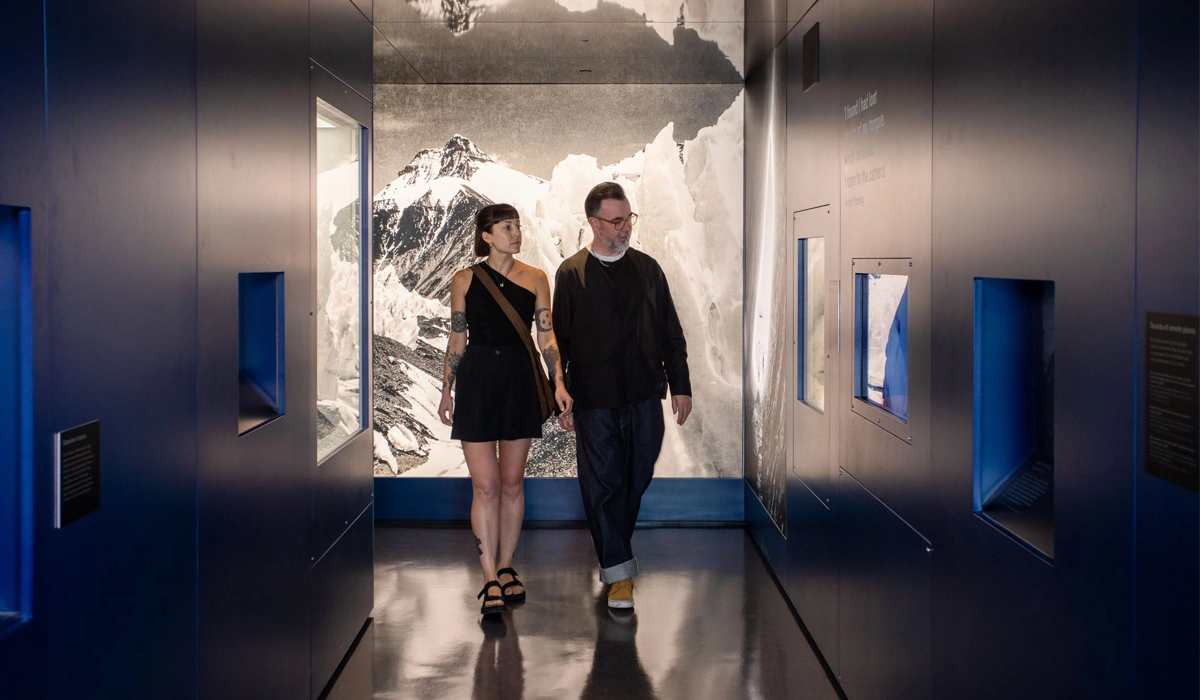 A couple walks through a darkened exhibition corridor with blue walls and display insets. A dramatic black-and-white mountain landscape is featured on the back wall, giving a sense of depth and atmosphere. The space has a modern, immersive design.