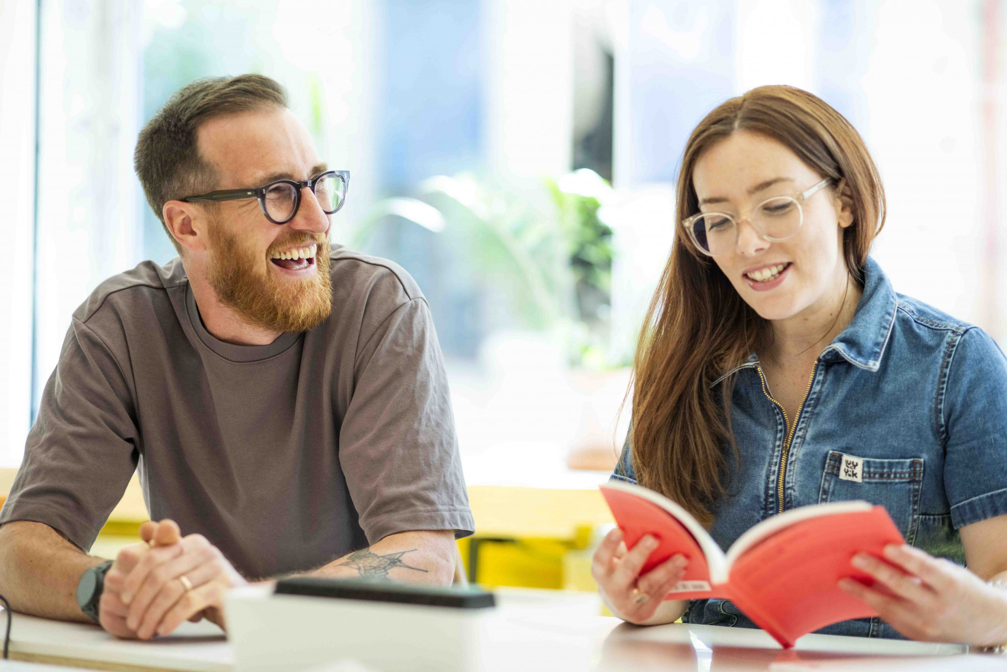 Two Mima team members sit at a table in a meeting room, Adam a white male with black glasses is laughing and Alice, a white female with clear rimmed glasses, is reading a book with a red cover.