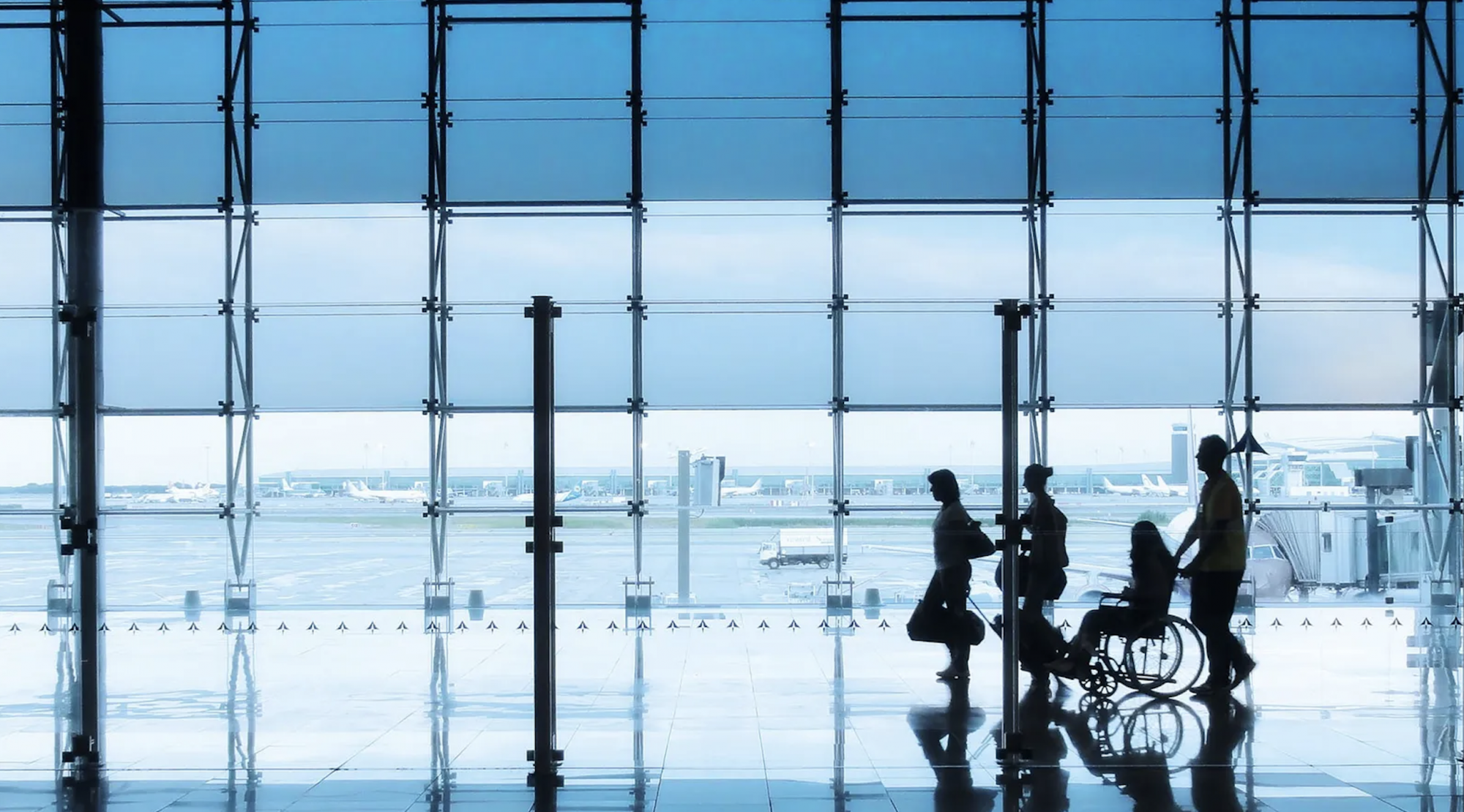 The silhouette of a small group of travellers - including a wheelchair user - move through an airport with planes and runways in the background