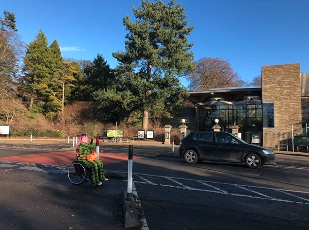 A photo of our Our Head of Accessibility and inclusive design, Emily Yates seated in her wheelchair outside the entrance to Royal Botanics Gardens Edinburgh