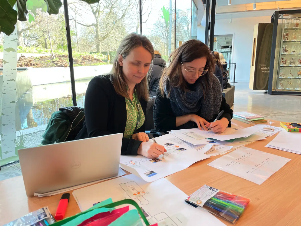 Two members of the Mima team are seated at a table in the Royal Botanic Gardens Edinburgh, manually mapping the visitor journey using pens and paper to map how visitors move through the space and their behaviour patterns and typical journeys.