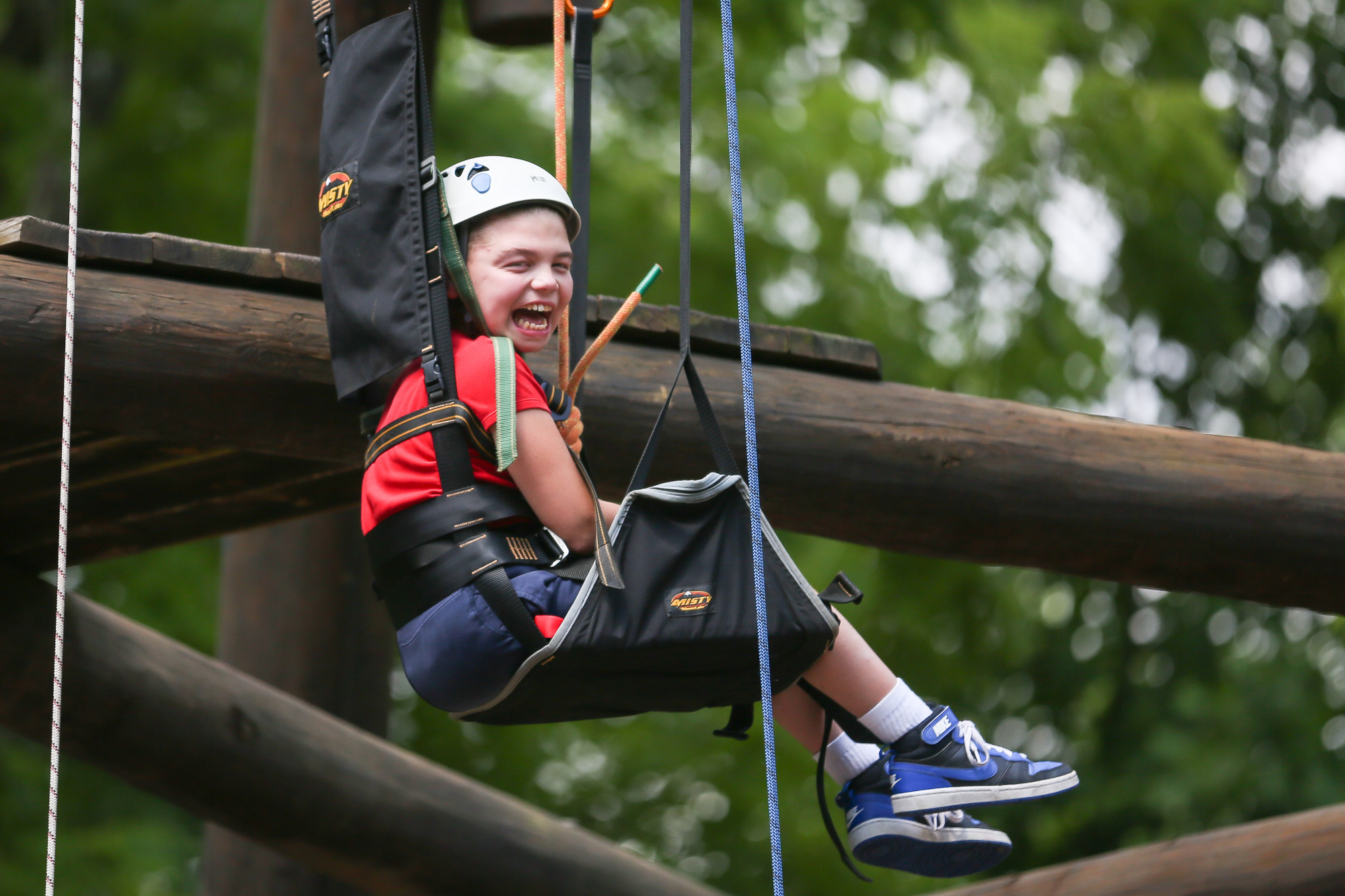 A picture of a smiling child in a specialist hoist surrounded by a wooden climbing structure amongst the trees