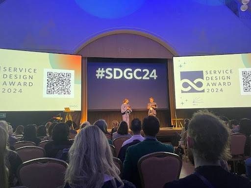 A photo of the main stage at Service Design Global Conference 2024 with two speakers on the stage holding microphones set against the backdrop of a videowall featuring three large screens