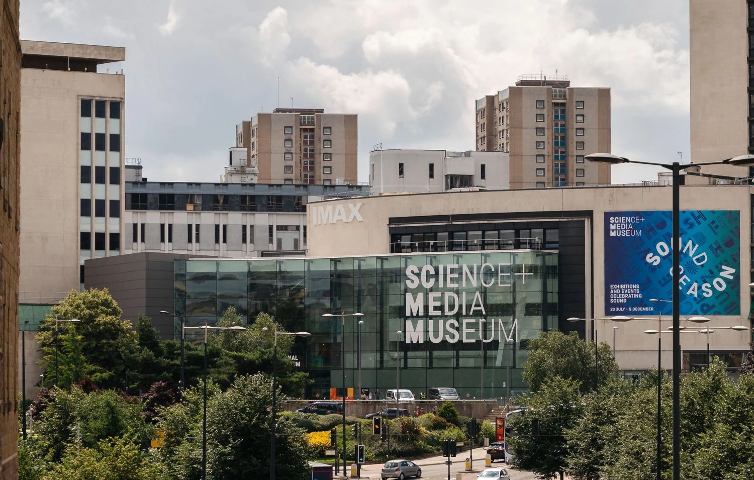 The image shows the exterior and entrance of the National Science and Media museum in Bradford, a contemporary building with high rise buildings behind it. A road and trees surrounds the front of the building.
