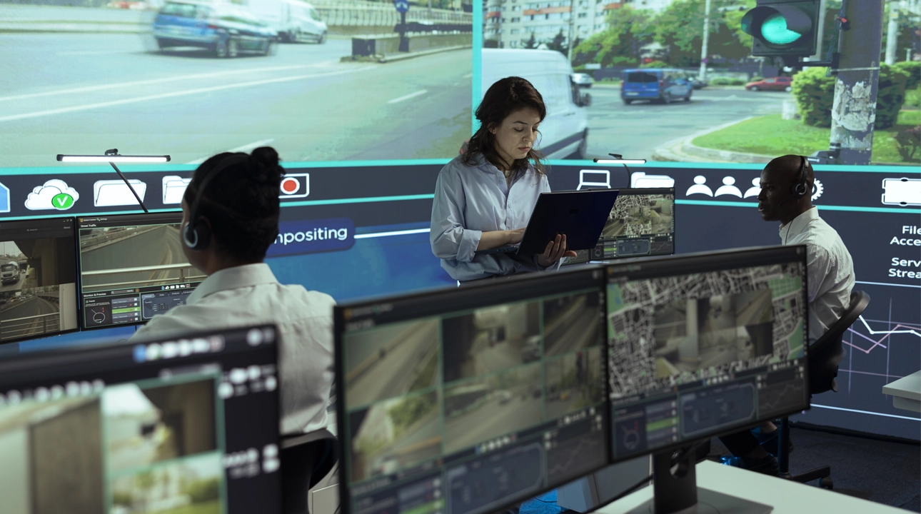 Team of control centre employees in a control room looking at screens with cars in traffic, monitoring urban activity in the city via CCTV surveillance footage on security cameras.