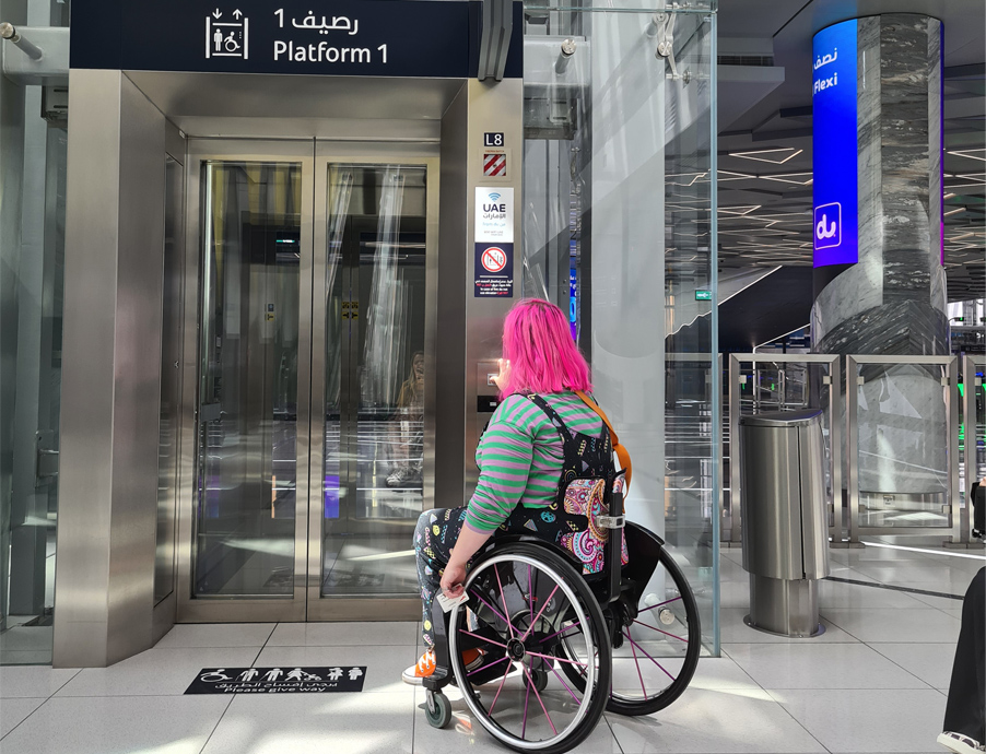 Emily, (Head of Accessibility and Inclusive Design at Mima) on site at COP28 conducting an accessibility audit, Emily, who is a wheelchair user, is outside a lift with signage directing to a platform.