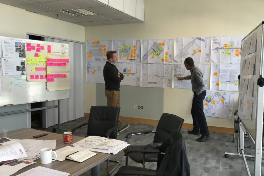 David, Mima's Managing Director, is white with blonde hair. He stands with a colleague in front of three workshopping walls - full of paper and colourful post-it notes. They are in discussion, with the colleague pointing to a particular detail on one of the pages.