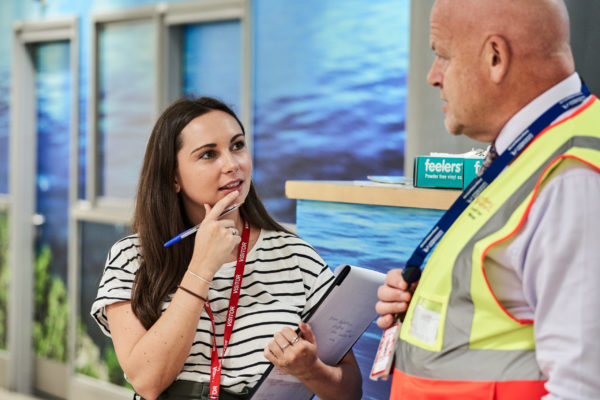 Ciara, a Mima team member, has dark hair and wears a black and white striped tshirt. She holds a pen to her mouth and looks contemplative whilst talking to a bald staff member, who wears a yellow hi-vis jacket.