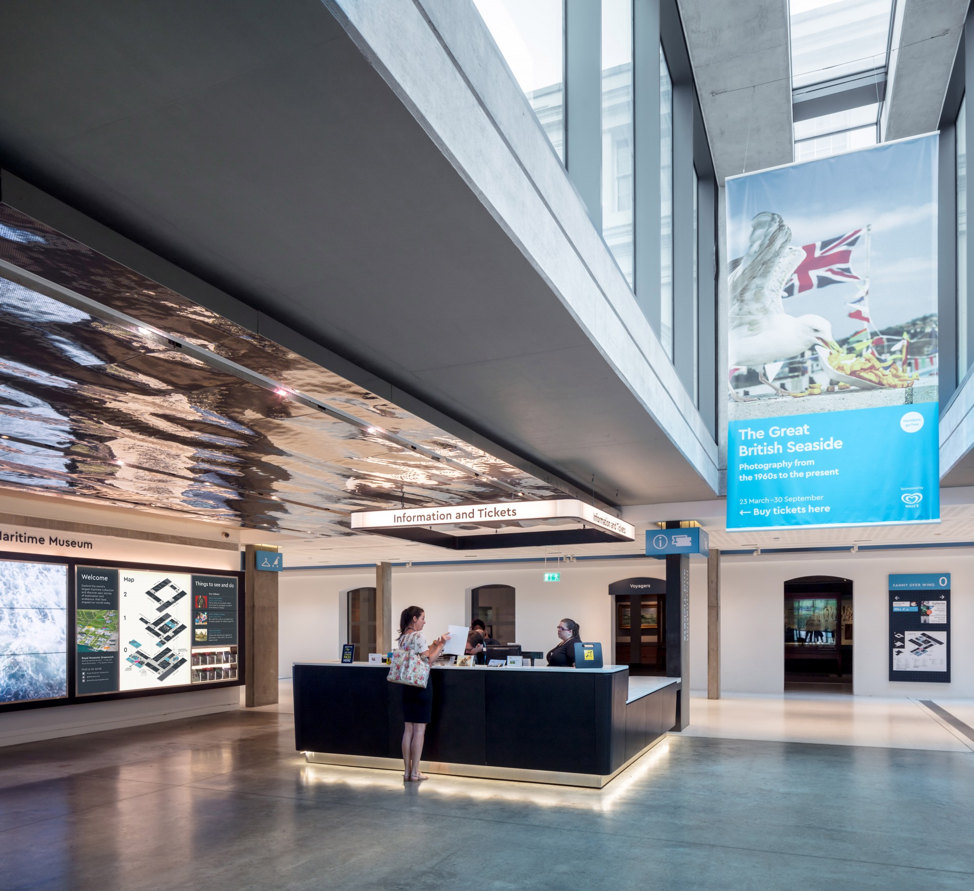 Inside the lobby of the National Maritime Museum. A square, black desk sits in the middle of the room and a member of staff is assisting a visitor in the distance. Maps of the museum floors are to the left, with a banner advertising 'The Great British Seaside' exhibition to the right. All elements are industrial in feel.