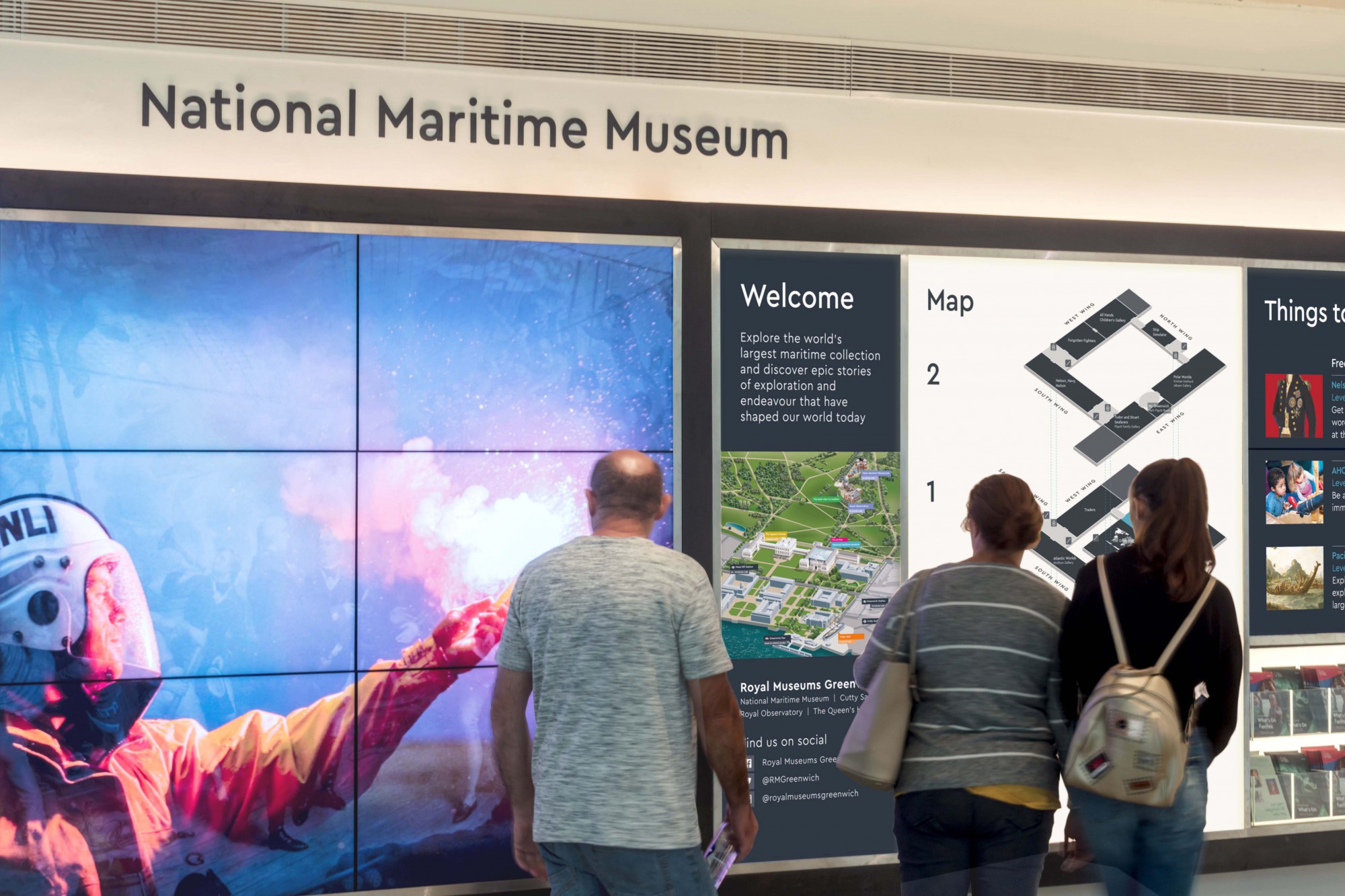 An image showing the welcome wall at the National Maritime Museum. The museum name is at the top of the white wall, in navy text, and a large video shows over multiple screens to the left. To the right, three visitors are standing facing the welcome map that Mima designed for the museum.