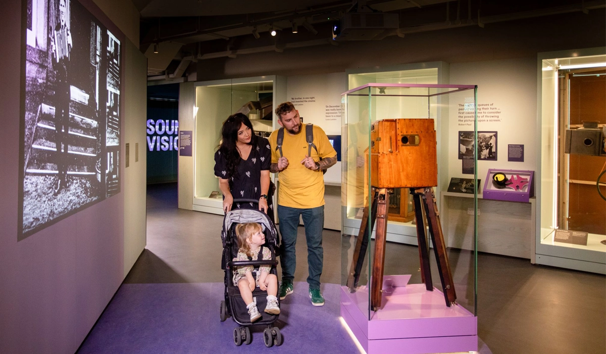 A young family explores a museum gallery featuring historic film and photography equipment. A woman pushes a child in a buggy while a man stands beside them, both looking at a large wooden early film camera displayed in a glass case. Behind them, a wall projection shows a black-and-white film image, and gallery text panels are visible around the room.
