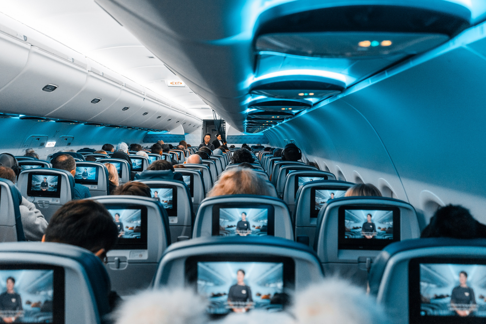 A picture of the inside of a full plane, with blue decor, as passengers watch a safety message.