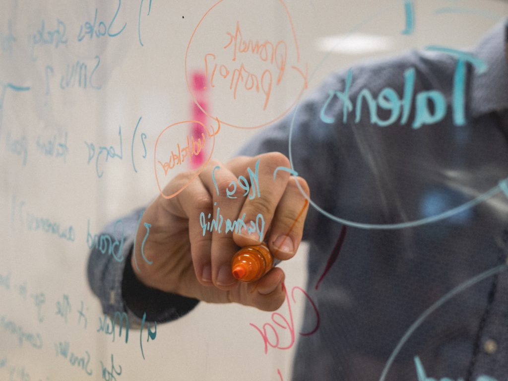 A clear board with colourful ideas written on it brings a workshopping atmosphere to the image. A black hand holds on orange pen, and is poised to add another thought onto the board.