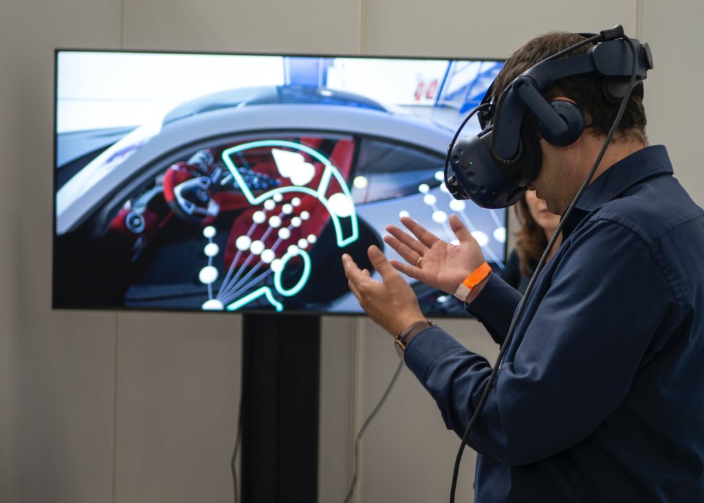 A white man with dark hair stands in front of a large monitor that is showing the red interior of a vehicle. The man wears a virtual reality headset, and is looking at his hands in disbelief.