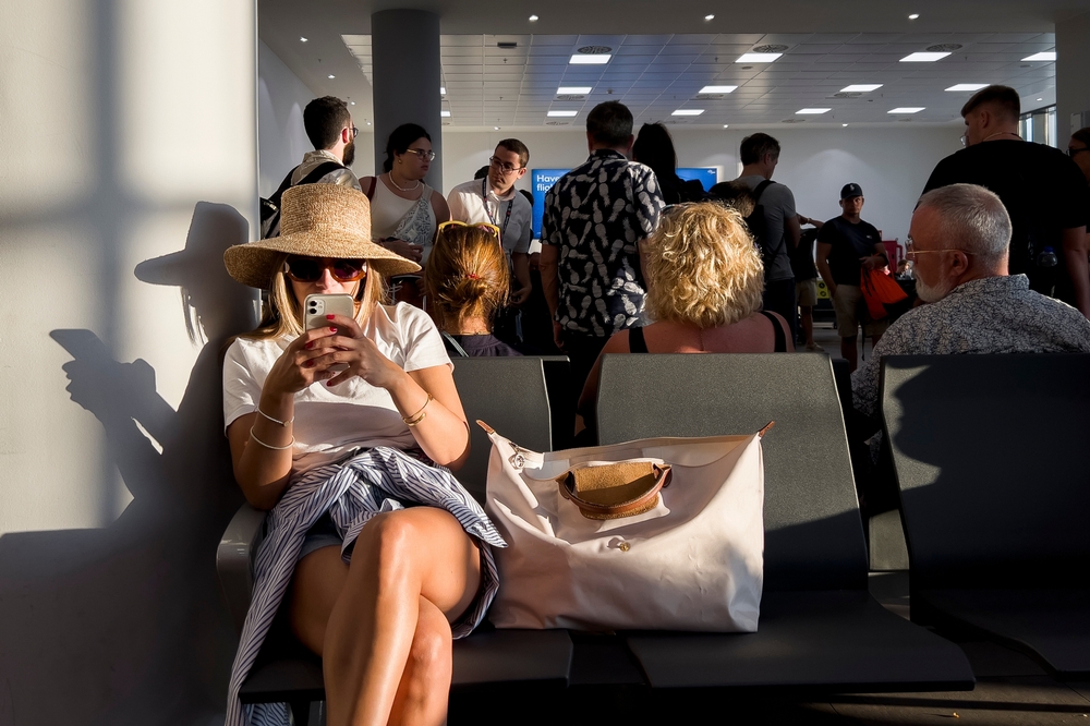 In the foreground a woman checks her phone with sunglasses and sunhat on, surrounded by passengers waiting for their flights in an international airport