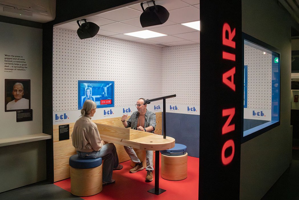 Two people sit inside a replica of a recording studio booth with the words 'on air' down the outside wall in red lettering, at the Sound + Vision galleries at the National Science museum.