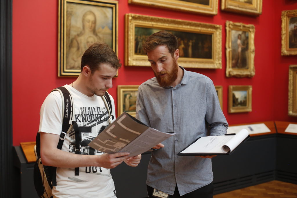 Adam, a Mima team member, is wearing a grey shirt and has a red beard. With crimson walls and traditional paintings behind him, he reads a museum map with a visitor, and holds a clipboard in one hand.