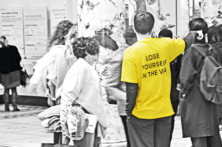 A black and white image of a staff member at the V&A gesticulating to direct a visitor. Both are facing away from the camera and only the backs of their bodies and heads are visible. In contrast to the black and white image, the staff member's t-shirt is bright yellow and has the phrase 'Lose yourself in the V&A' written on the back.