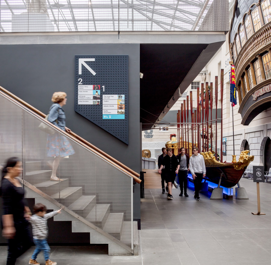 A photo of the finished in-situ wayfinding system in place at the National Maritime Museum Greenwich with a visitor walking down the stairs near the sign