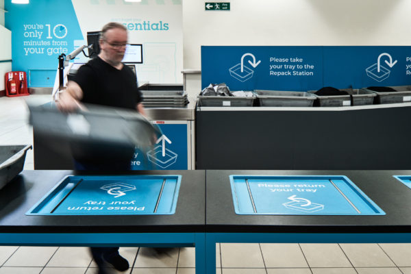 The security area at John Lennon Airport. A male passenger with grey hair, glasses and a black t-shirt picks up a luggage tray from a security station. Underneath the tray is a turquoise poster stating 'Please return your tray'.