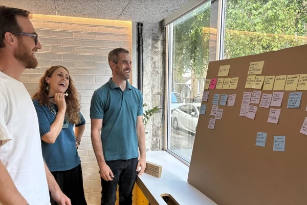 Three people stand in front of a large brown board with post it notes on. They are talking and writing notes on the board.
