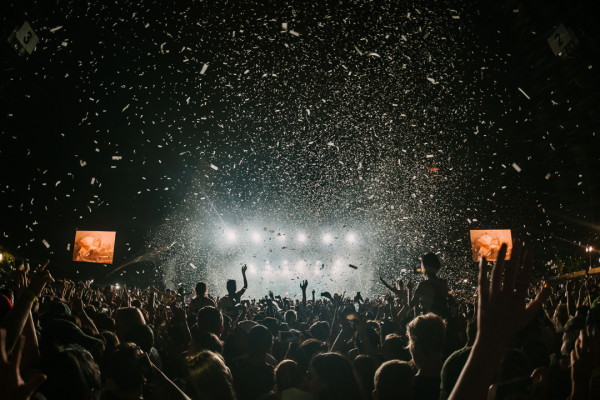 A large crowd of people is gathered at an outdoor concert at night. The stage is brightly lit with white lights beaming, and confetti is falling from above. Many attendees have their hands raised, and the scene captures the excitement of the event.