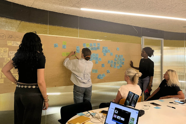 The Tellmi and Mima team conducting an ideation workshop in an office with glass walls. On the walls they are placing post it notes and writing comments on a large sheet of brown paper.